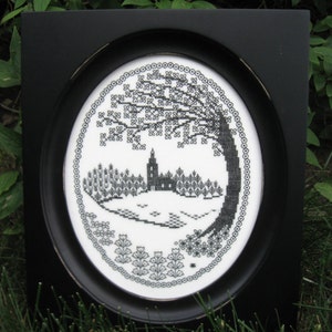 May include: A black oval frame containing a white fabric piece with a black cross-stitch embroidery design. The embroidery depicts a central church building surrounded by trees, a large tree on the right, and floral motifs at the bottom.