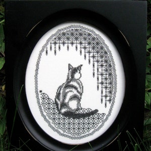 May include: A black and white cross-stitch embroidery featuring a cat sitting on a patterned surface, enclosed within an oval black frame. The design includes intricate black thread detailing against a white fabric background with a lace-like border.