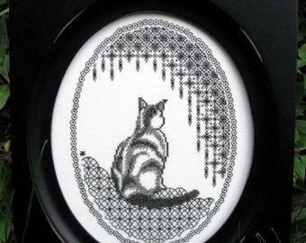 May include: Black and white embroidery of a cat sitting under a lacy design in a black oval frame.