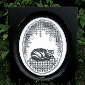 May include: A black oval frame containing a white fabric piece with a black cross-stitch embroidery of a sleeping cat. The cat rests on a patterned base, surrounded by an intricate black lace-like border that arches over the feline figure.