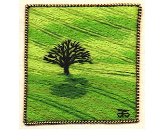 May include: Square embroidered artwork featuring a black tree on a green field. The artwork is framed with a gold-colored border. The background is a textured green, with the tree casting a shadow. The artist's name, Tanja Berlin, is at the bottom.