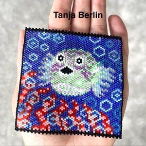 May include: A square beaded patch featuring a pixelated pufferfish design in shades of green, purple, and white against a deep blue background with red coral-like accents. The text Tanja Berlin is printed in black above the square beaded piece.