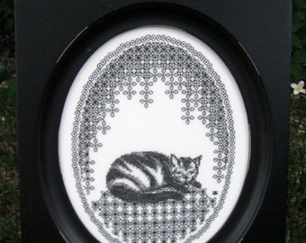 May include: Black and white embroidery of a cat sleeping in a circular frame with a black border. The embroidery is surrounded by a black and white geometric pattern.
