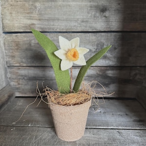 May include: A handcrafted felt daffodil in a small burlap pot. The white flower has a yellow center, with green leaves. The pot is filled with brown straw. The background is a wooden surface.