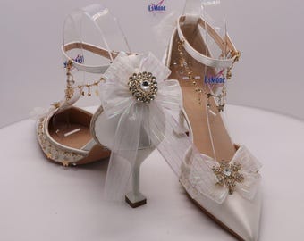 Könnte beinhalten: Weiße Satin-High Heels mit spitzer Zehenpartie und Knöchelriemen. Die Absätze verfügen über dekorative Schleifen mit Kristallverzierungen und baumelnden Charms. Die Schuhe haben ein zartes, elegantes Design, passend für besondere Anlässe.
