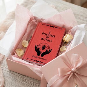 May include: A red hardcover book titled A Discovery of Witches by Deborh Harkness, featuring a black graphic of hands holding a crystal ball, nestled in a light pink gift box lined with white tissue paper, dried flowers, and a pink ribbon bow.