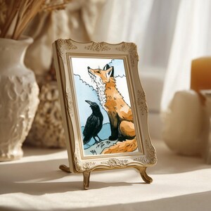 May include: A rectangular picture frame with an ornate, cream-colored border featuring embossed floral details. Inside the frame is a watercolor illustration of a ginger fox sitting next to a black crow against a light blue sky background.