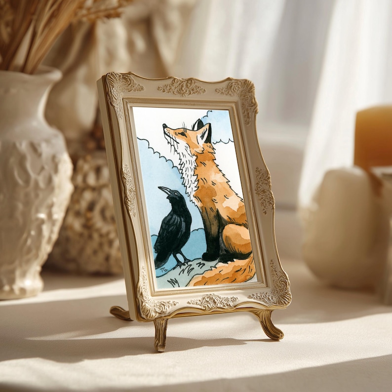 May include: A rectangular picture frame with an ornate, cream-colored border featuring embossed floral details. Inside the frame is a watercolor illustration of a ginger fox sitting next to a black crow against a light blue sky background.