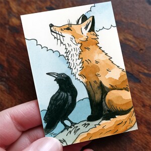 May include: A rectangular art print featuring a watercolor illustration of a seated orange fox with a white chest and a black raven standing beside it. The background shows stylized light blue clouds and the initials TG are visible in the corner.