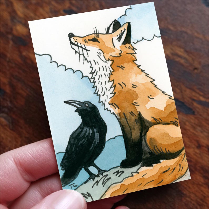 May include: A rectangular art print featuring a watercolor illustration of a seated orange fox with a white chest and a black raven standing beside it. The background shows stylized light blue clouds and the initials TG are visible in the corner.