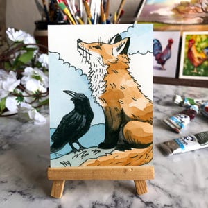 May include: A watercolor painting on a small rectangular card displayed on a wooden easel. The artwork depicts a seated orange fox looking upward next to a black crow against a light blue sky with white clouds. The initials TG are visible.