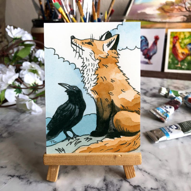 May include: A watercolor painting on a small rectangular card displayed on a wooden easel. The artwork depicts a seated orange fox looking upward next to a black crow against a light blue sky with white clouds. The initials TG are visible.