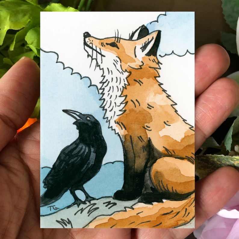 May include: A watercolor illustration on a rectangular card featuring a seated orange and white fox looking upward next to a black crow. The background consists of light blue sky and white clouds. The initials TG are visible in the bottom left.