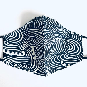 May include: A navy blue and white patterned face mask with black ear loops. The mask features a repeating wave-like design in white against a dark blue background. The mask is made of fabric and is designed to cover the nose and mouth.