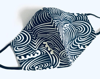 May include: A navy blue face mask with a white wave pattern. The mask has black elastic ear loops and is made of fabric. The design features swirling lines and curves, creating a dynamic and stylish look. This reusable mask is suitable for everyday wear.