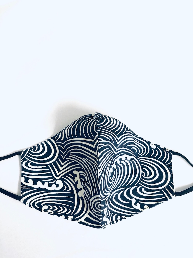 May include: A navy blue and white patterned face mask with black ear loops. The mask features a repeating wave-like design in white against a dark blue background. The mask is made of fabric and is designed to cover the nose and mouth.