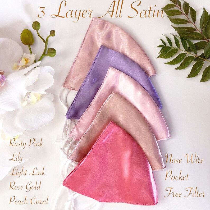 Puede incluir: Cinco mascarillas de sat&eacute;n en diferentes tonos de rosa, morado y coral. Las mascarillas est&aacute;n apiladas, con la mascarilla superior de color rosa claro. El texto "3 Layer All Satin" est&aacute; en la parte superior de la imagen. El texto "Rusty Pink Lily", "Light Link", "Rose Gold" y "Peach Coral" est&aacute; listado debajo de las mascarillas. El texto "Nose Wire", "Pocket" y "Free Filter" est&aacute; listado en el lado derecho de la imagen.