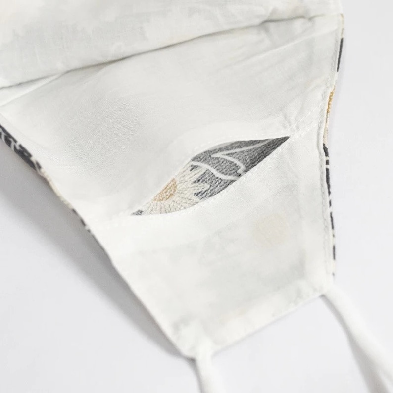 May include: White face mask with a floral pattern. The mask has a pocket and white ties. The floral pattern includes daisies and leaves in black, white, and gold.