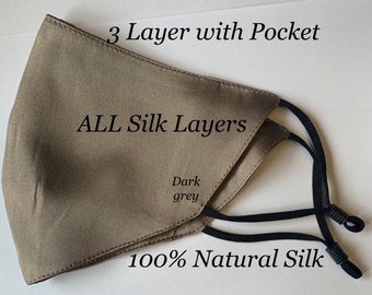 Op de afbeelding: Een donkergrijze zijden gezichtsmasker met een zakje. Het masker is gemaakt van 100% natuurlijke zijde en heeft drie lagen. De tekst "3 Layer with Pocket", "ALL Silk Layers", "Dark grey" en "100% Natural Silk" staat op het masker gedrukt.