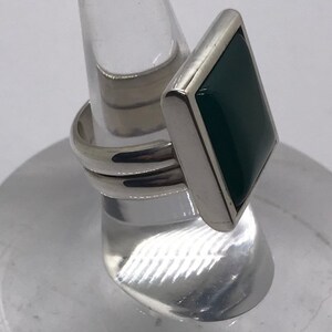 Op de afbeelding: Een zilveren ring met een vierkante groene steen gezet in een zilveren bezel. De ring heeft een brede band met een textuur.
