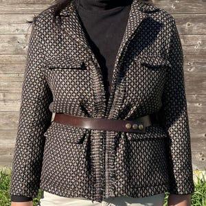 Peut inclure: Une veste en tweed noir et blanc avec une ceinture en cuir marron et des accents dorés. La veste a une bordure effilochée et deux grandes poches sur le devant.