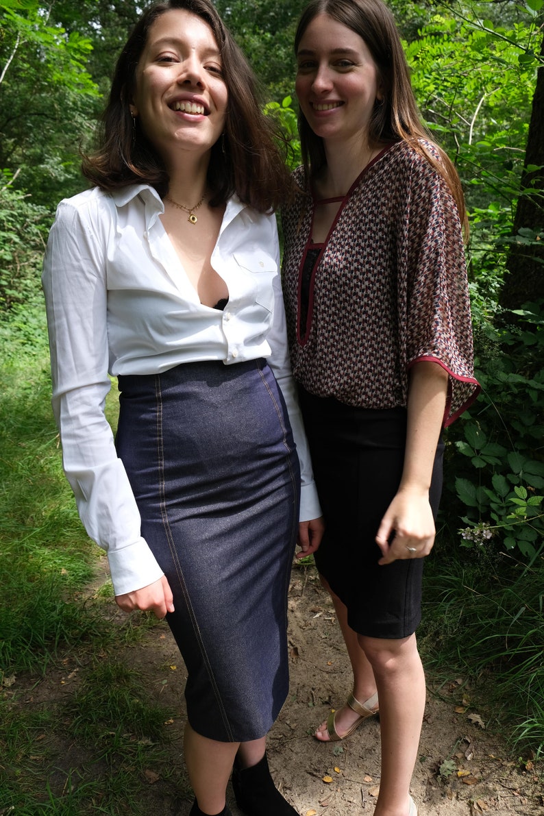 May include: Two women standing in a forest, one wearing a white button-down shirt with a denim skirt and the other wearing a patterned blouse with a black skirt.