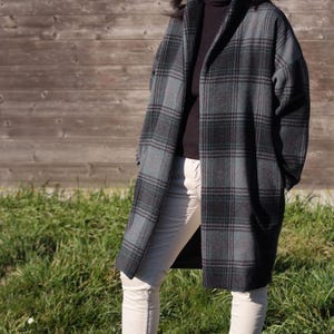 Peut inclure: Une femme portant un long manteau à carreaux gris et noir avec un pull à col roulé noir et un pantalon blanc. Elle porte des lunettes de soleil et a les cheveux foncés.