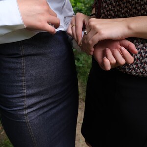 May include: Two people are standing next to each other, holding hands. The person on the left is wearing a white shirt and a dark blue denim skirt. The person on the right is wearing a black and white patterned blouse and a black skirt.
