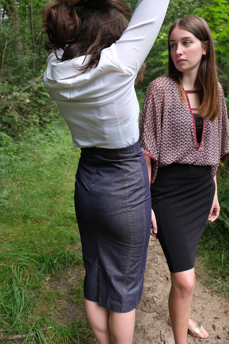 May include: Two women wearing skirts stand in a forest. The woman in the foreground is wearing a white button-down shirt and a dark blue denim skirt. The woman in the background is wearing a patterned blouse and a black skirt.