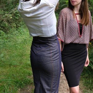 May include: Two women wearing skirts stand in a forest. The woman in the foreground is wearing a white button-down shirt and a dark blue denim skirt. The woman in the background is wearing a patterned blouse and a black skirt.