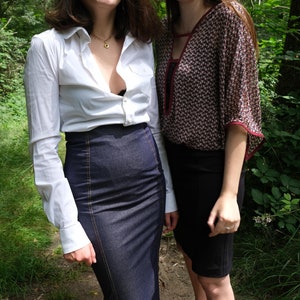 May include: Two women standing in a forest, one wearing a white button-down shirt with a denim skirt and the other wearing a patterned blouse with a black skirt.