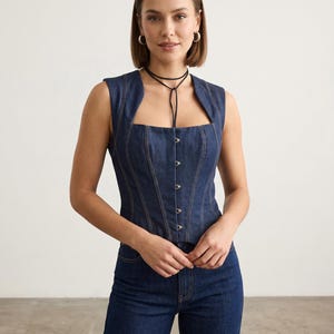 Peut inclure: Un haut corset en jean foncé sans manches, avec un col carré et une fermeture boutonnée. Il est associé à un jean en denim foncé. La personne porte un ras du cou noir et des créoles argentées. L'ensemble est moderne.