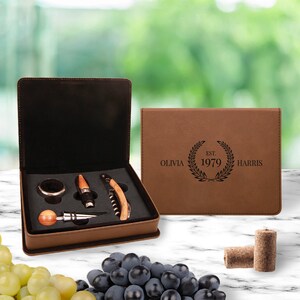 May include: A brown leatherette gift box with a wine opener set inside. The box is engraved with the text "Olivia EST. 1979 HARRIS". The set includes a bottle opener, a stopper, a ring, and a foil cutter.