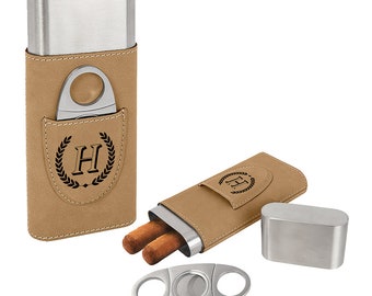 May include: A brown leather cigar case with a silver metal insert and a built-in cigar cutter. The case has a monogrammed "H" with a laurel wreath design. Two cigars are visible in the case.