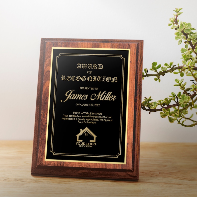 May include: A brown wooden plaque with a gold trim and a black background. The plaque has a gold inscription that reads "Award of Recognition" presented to "James Miller" on August 27, 2022. The inscription also includes the text "Most Notable Patron" and a small house logo with the text "Your Logo" and "Slogan Here".