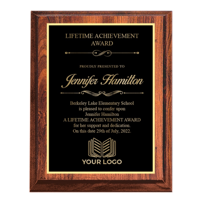 May include: A black plaque with gold lettering on a wooden frame. The plaque reads "Lifetime Achievement Award" and is presented to Jennifer Hamilton. The plaque also states that Berkeley Lake Elementary School is pleased to confer upon Jennifer Hamilton a Lifetime Achievement Award for her support and dedication on this date 29th of July, 2022. The plaque has a book logo with the text "Your Logo" underneath.