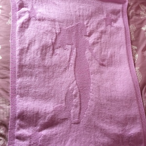 May include: A lilac-colored knitted blanket featuring a silhouette of a cactus and several star shapes. The blanket has a ribbed border and is made of soft yarn. The cactus design is in the center, with stars in each corner.