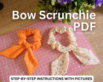 May include: Two fabric scrunchies with bows. One scrunchie is orange with white polka dots and the other is white with small yellow flowers. The scrunchies are on a pink cutting mat with white grid lines. The text "Bow Scrunchie PDF" is at the top of the image.