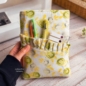 May include: A fabric pouch featuring a white background with a pattern of green and yellow floral motifs. The pouch has a ruffled front pocket containing two pens and a small clear plastic ruler. The text craftsonfire/hanasews is visible.
