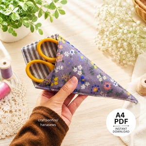 Puede incluir: Una funda triangular de tela para tijeras con un exterior morado con un patrón floral multicolor y un forro interior a rayas grises y blancas. La funda contiene unas tijeras con mangos amarillos. El texto en la imagen dice A4 PDF.