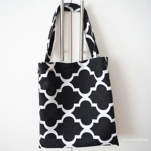 May include: A black and white tote bag featuring a geometric quatrefoil design. The bag has two handles and is suspended against a white backdrop. The bag is made of fabric and is likely used for carrying items.