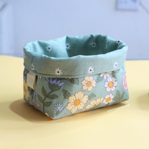 May include: A light green fabric storage basket with a floral pattern featuring white, yellow, and blue flowers. The basket has a white label with the text "CRAFTSONFIRE.COM".