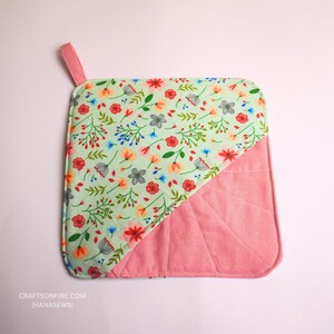 Könnte beinhalten: Quadratischer, gesteppter Topflappen mit einer rosa Schlaufe. Die obere linke Ecke hat ein Blumenmuster auf hellgrünem Hintergrund, während die untere rechte Ecke einfarbig rosa ist. Der Topflappen ist ca. 20 cm groß.