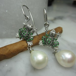 May include: Silver dangle earrings with a green frog charm and a white pearl drop.