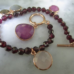 May include: A garnet bead bracelet with a toggle clasp. The bracelet has three charms: a pink, a gray, and a green gemstone.
