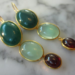 May include: Gold-toned dangle earrings featuring three oval-shaped stones in shades of green and burgundy. The stones are set in gold bezels, creating a striking contrast. The earrings have a classic, elegant design.