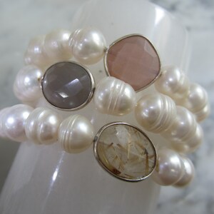 May include: A pearl bracelet with three different colored gemstone accents. The bracelet is made of white pearls and features a light pink, gray, and clear gemstone.