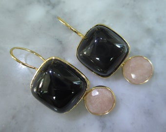May include: A pair of gold-coloured drop earrings. Each earring features a large, square, black stone with a gold border, paired with a smaller, round, light pink stone. The earrings have a simple hook design.