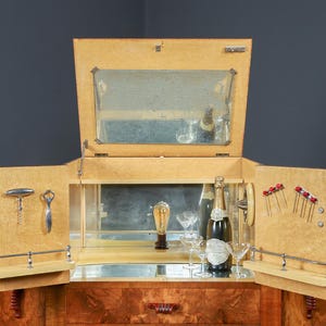 May include: A vintage wooden bar cabinet with mirrored interior and open doors. The cabinet features a bottle of champagne, glassware, and bar tools. The cabinet is made of light-colored wood with a dark wood base.