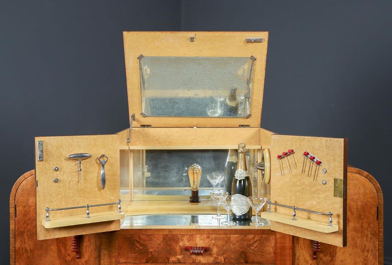 May include: A vintage wooden bar cabinet with mirrored interior and open doors. The cabinet features a bottle of champagne, glassware, and bar tools. The cabinet is made of light-colored wood with a dark wood base.